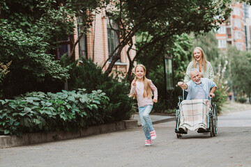 Happy family outing with granddaughter running beside grandfather in a wheelchair pushed by mother outdoors in a green city park