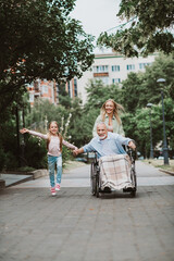 Fototapeta premium Joyful family outing a grandfather in a wheelchair being pushed by his daughter and a smiling girl running in a park