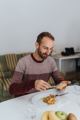 A man is enjoying a casual meal at home, spreading butter on bread, with a light setting and fresh ingredients.