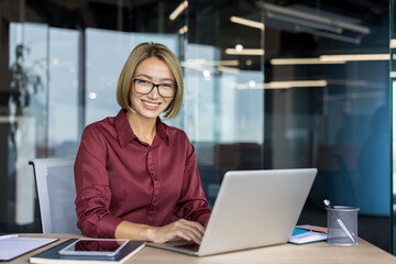Businesswoman working on a laptop, sitting at her desk in a contemporary office setting, smiling at...