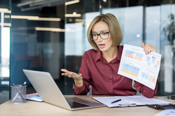 Businesswoman sitting at desk in modern office, having a virtual meeting on laptop, holding up financial charts and reports, and discussing data with a serious expression