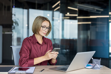 Businesswoman with glasses sitting at an office desk, actively using a smartphone while a laptop is open beside her, representing modern communication and remote work