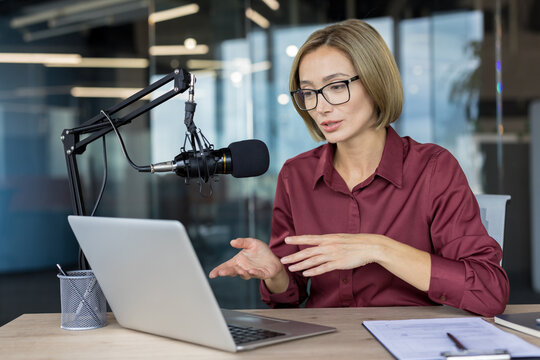 Woman in glasses talking into a professional microphone, broadcasting a podcast or online webinar from her laptop in a modern office setup, creating digital content
