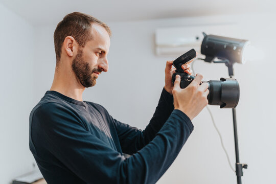 A man prepares his camera equipment indoors, adjusting settings on a digital camera with focus lighting placed nearby, showcasing professional photography session preparations.