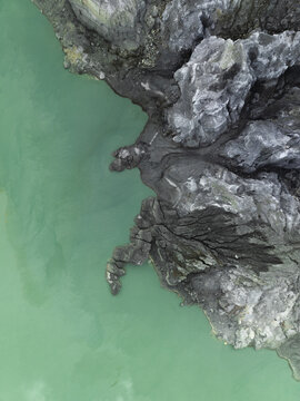 Aerial view of jagged, dark rock formations meet the pale green waters of Ijen Crater Lake, creating a striking contrast of textures and tones, Mount Ijen, East Java, Indonesia.
