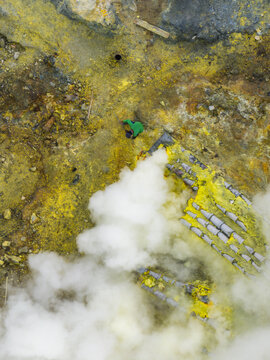 Aerial view of a miner extracting sulfur amidst vibrant yellows and grays, with plumes of white smoke rising from the volcanic landscape, Mount Ijen, East Java, Indonesia.