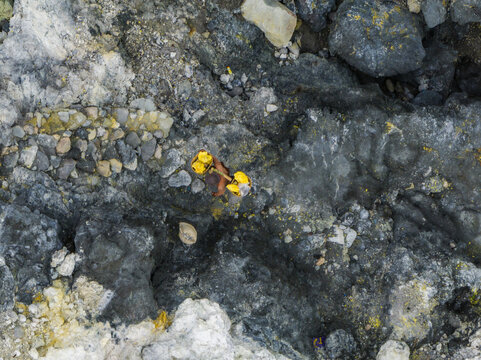 Aerial view of the rugged, textured landscape reveals a tapestry of greys, yellows, and whites, etched with mineral deposits and rocky formations, Mount Ijen, East Java, Indonesia.