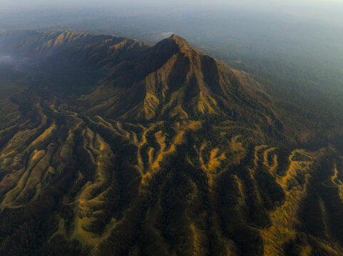 Aerial view of the rugged Ijen volcano's peak rises majestically amidst a tapestry of verdant forests and golden grasslands, Mount Ijen, East Java, Indonesia.