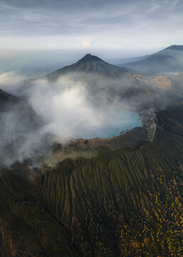 Aerial view of the Ijen volcano's turquoise crater lake steaming amidst the rugged, verdant slopes, shrouded in ethereal mist, Mount Ijen, East Java, Indonesia.