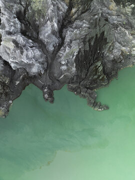 Aerial view of stark, grey cliffs meet the surreal, turquoise waters of the volcanic lake, creating a dramatic contrast of textures and tones, Mount Ijen, East Java, Indonesia.