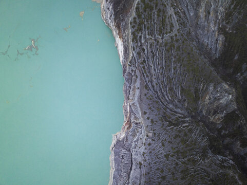 Aerial view of the turquoise lake contrasting with the rugged, textured cliffs of Mount Ijen, East Java, Indonesia.