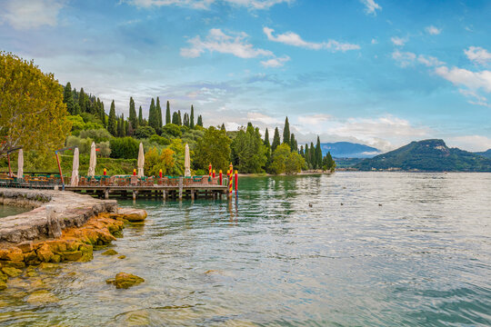 Porto di Punta San Vigilio on a sunny day, showcasing sparkling waters, colorful boats, charming lakeside buildings, and the surrounding lush greenery under blue skies — a picturesque Lake Garda scene