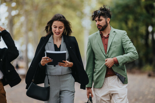 Two people walk along a city path on a fall day. A woman in a gray suit holds a notebook while a man in a green blazer accompanies her, sharing a casual business moment.