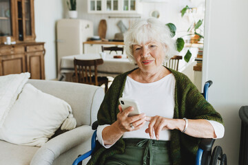 Adorable disabled elderly woman in wheelchair using smartphone sitting in cozy living-room of...
