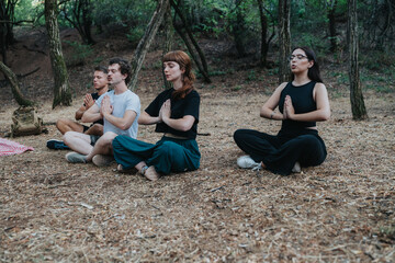 A diverse group sits cross legged on a forest floor with hands in a prayer pose, surrounded by trees. They share a moment of calm, focus, and mindfulness in nature.