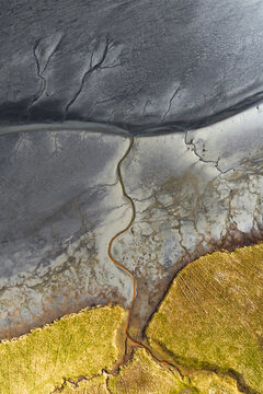 Aerial view of winding waterways carve through a tapestry of textures, where stark grey earth meets golden fields in a mesmerising dance of nature's artistry, South Coast, Iceland.