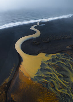 Aerial view of a winding river snaking through black sands towards the ocean, where foamy waves crash under a misty sky, South Coast, Iceland.