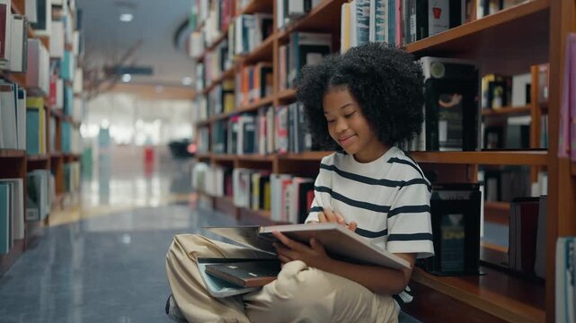 American elementary student sitting on the floor in the library while studying and reading a book. Education, Learning and knowledge concept