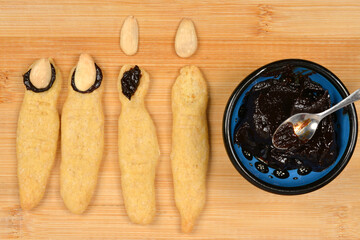An overhead shot showing the process of decorating baked witch finger cookies by attaching almond nails with dark plum jam, next to a bowl of jam