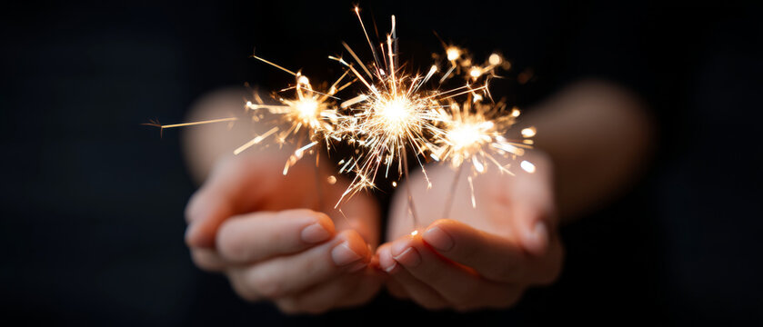 Close-up of hands holding bright sparklers glowing against a dark background with sparks flying in multiple directions