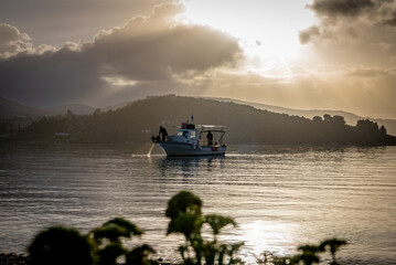 Fototapeta premium The Autumn sun breaking through the clouds casting sun rays on the still sea with a small fishing boat and fisherman checking their catch.