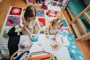 Two young girls in a vibrant playroom create artwork using drawing tools, surrounded by art supplies and a playful interior. A cheerful moment of childhood creativity and bonding.