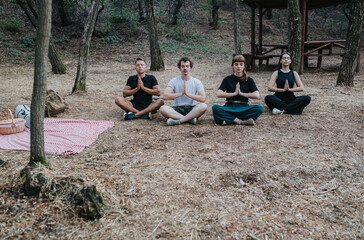 A group of four people sits cross-legged in a tranquil forest, hands in prayer. A red-and-white picnic blanket and baskets rest nearby, suggesting a peaceful outdoor retreat.