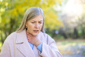 Senior woman clutching her throat in pain, looking unwell in a chilly autumn park, showing symptoms of sore throat, cold or seasonal flu and discomfort