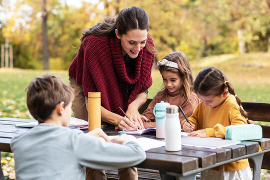 The teacher and children are now sitting together at a wooden table in the park, talking and observing nature around them. The moment is calm and focused, showing a gentle learning atmosphere outdoors
