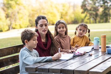 The teacher and children are now sitting together at a wooden table in the park, talking and observing nature around them. The moment is calm and focused, showing a gentle learning atmosphere outdoors