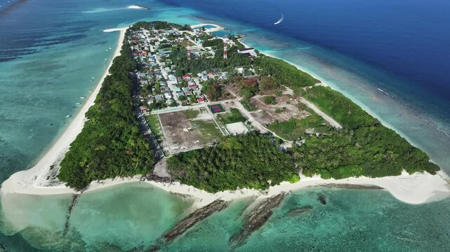 Aerial view of an island showcasing vibrant turquoise waters meeting sandy shores and lush green vegetation contrasting with the built environment, Naavaidhoo, Haa Dhaalu City, Maldives.