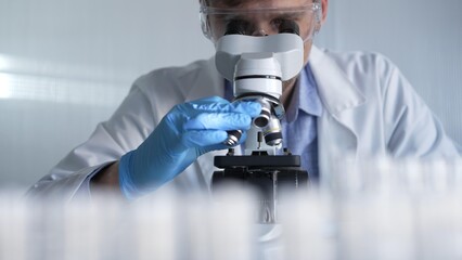 Male scientist researcher wearing blue gloves, protective glasses is using microscope in modern...