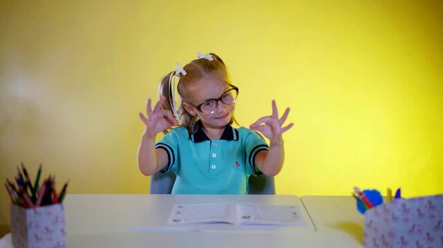Elementary-aged girl seated at a desk with art supplies, displaying an OK sign. Young student with pigtails and glasses making "okay" signs against a vibrant yellow background.