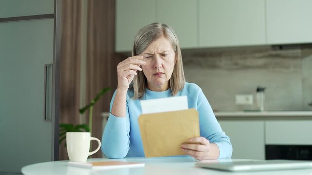 Disappointed senior gray haired female reading letter with bad news sitting in kitchen at home. Sad elderly retired woman is depressed by an unpleasant notification, shocked by information in document