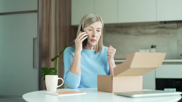 Unhappy senior female arguing on the phone after receiving a parcel with the wrong product at home. Elderly woman makes a bad review, she is dissatisfied with shopping in an online store. Damaged item