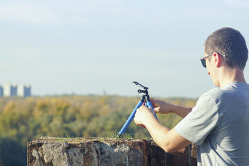 A tourist standing at a viewpoint sets up a portable tripod to film himself with a smartphone. Concept of self-recording exploration, autonomous travel experience, and personal storytelling in motion.