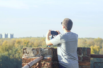 Fototapeta premium Man mounting smartphone on octopus tripod at scenic overlook with city horizon. Represents timelapse recording setup, hands-free filming solution, travel vlogging equipment, and mobile journalism tool