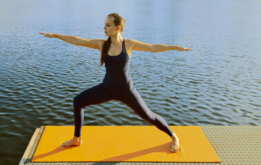 Middle aged young adult latino hispanic lady female girl doing warrior yoga pose by the sea on water surface background. woman standing barefoot on pier doing virabhadrasana on lake river shore