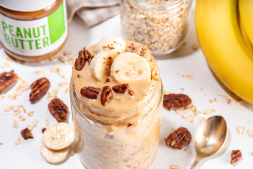 Peanut butter and banana overnight oatmeal, tasty sweet breakfast overnight oats in jar, with ingredients for cooking on kitchen table