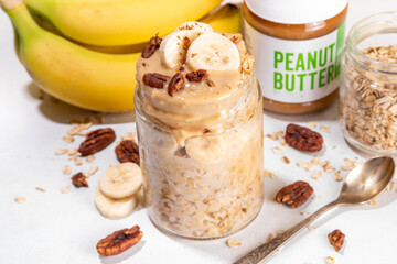Peanut butter and banana overnight oatmeal, tasty sweet breakfast overnight oats in jar, with ingredients for cooking on kitchen table