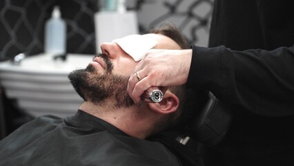 Professional barber grooming client's beard using electric razor, delivering precise trimming in...