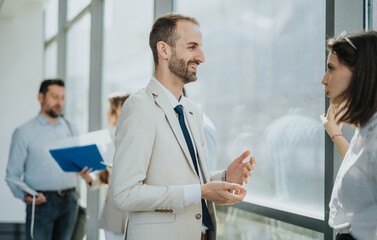Fototapeta premium A smiling professional in a light suit chats with a colleague near a large window while others review documents in the background, capturing workplace collaboration and networking.