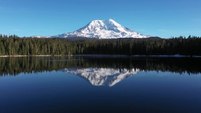 4K aerial drone footage of Takhlakh Lake and snow covered Mount Adams in Washington State USA during late fall early winter. Scenic reflection of mountain in calm water surrounded by evergreen forest 