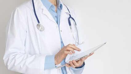Unknown female doctor, wearing white coat over a blue blouse and a stethoscope, is using a digital tablet, suggesting innovative healthcare practices in medicine. Health care and medicine concept