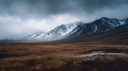 Moody mountain landscape with snow covered peaks and expansive brown grass field under a cloudy dramatic sky