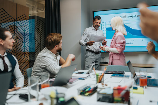 A diverse group of professionals collaborates around a table, reviewing papers and notes as a presenter discusses data on a large screen in a bright conference room.