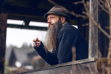 Bearded man in traditional hat smoking a pipe on wooden balcony of rustic cabin, peaceful rural lifestyle in mountain region