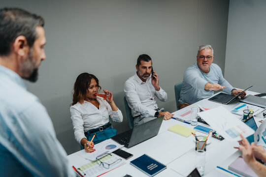 A group of professionals sits around a conference table, sharing ideas, reviewing charts, and coordinating tasks. Laptops, notes, and documents reflect a collaborative business planning session.