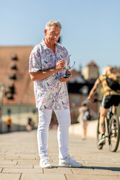 Confident best ager tourist in floral shirt and white pants standing on street in Regensburg, Germany on a sunny summer day