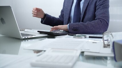 Businessman wearing blue suit is working with laptop while sitting and gesturing with his hands at the glass table in office background. Low lighting, close-up view. Business people and audit concept
